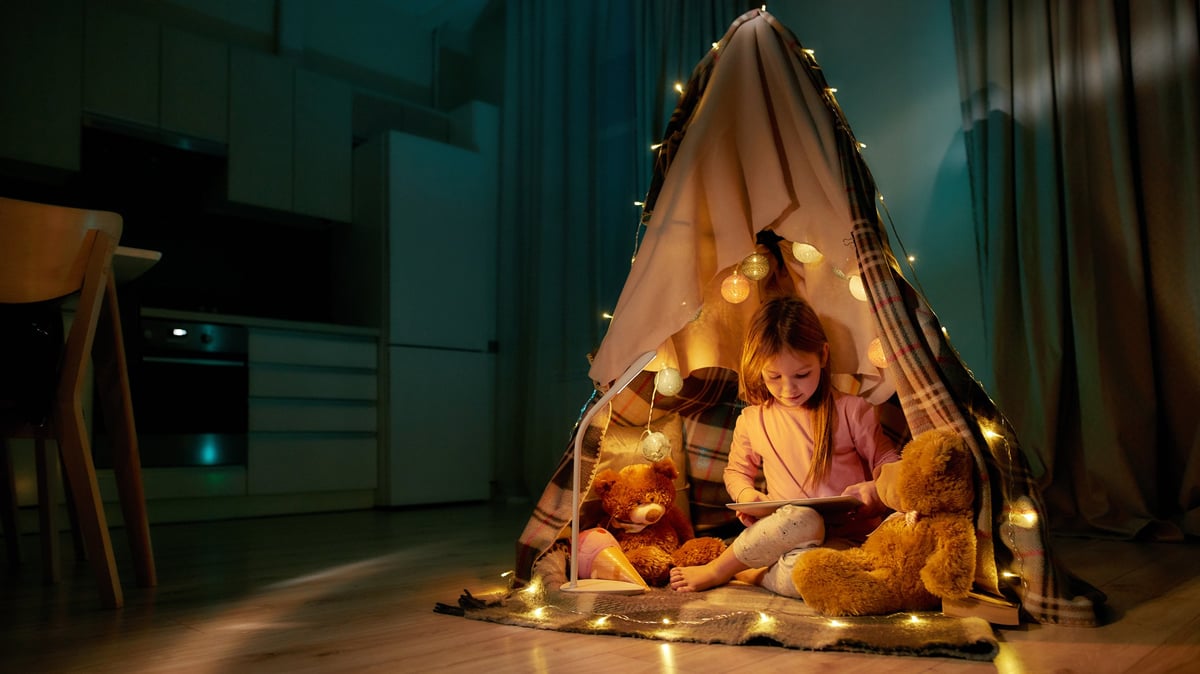 A little girl in pajamas in a cozy play tent with fairy lights and stuffed animals