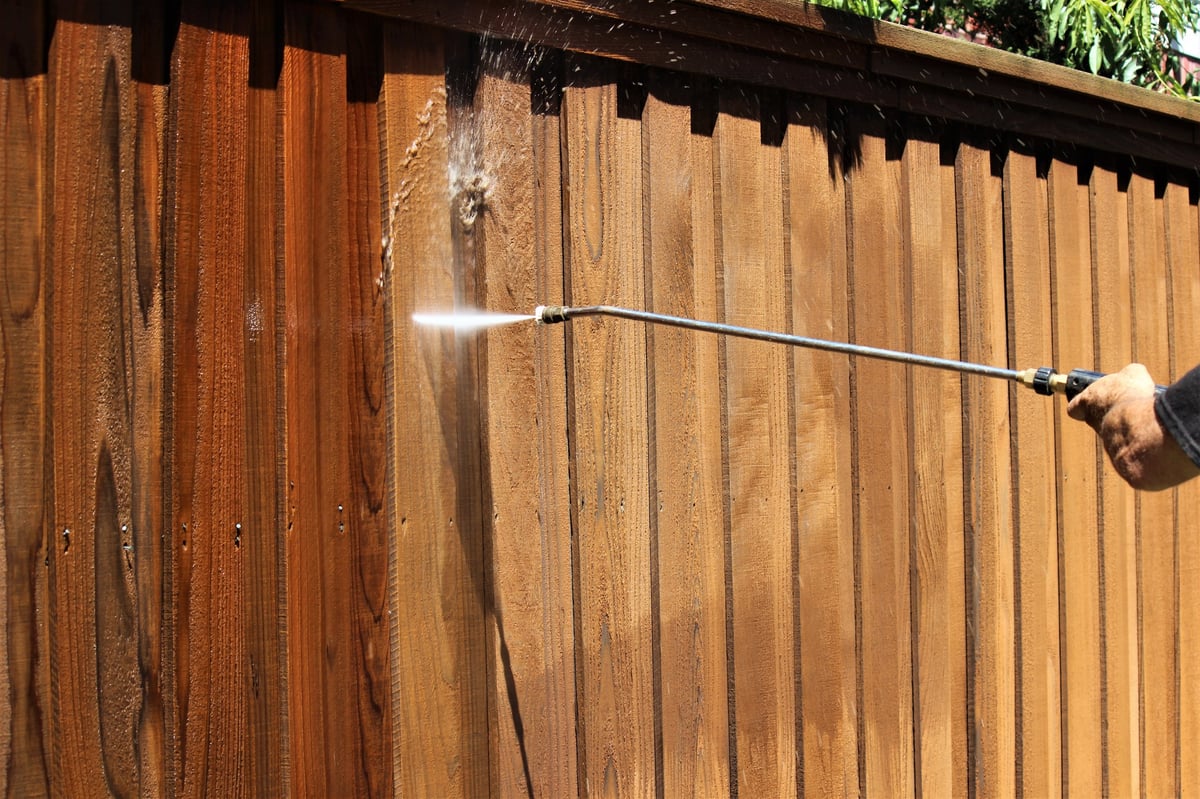 Worker cleaning a wooden fence with a pressure washer, removing dirt and algae