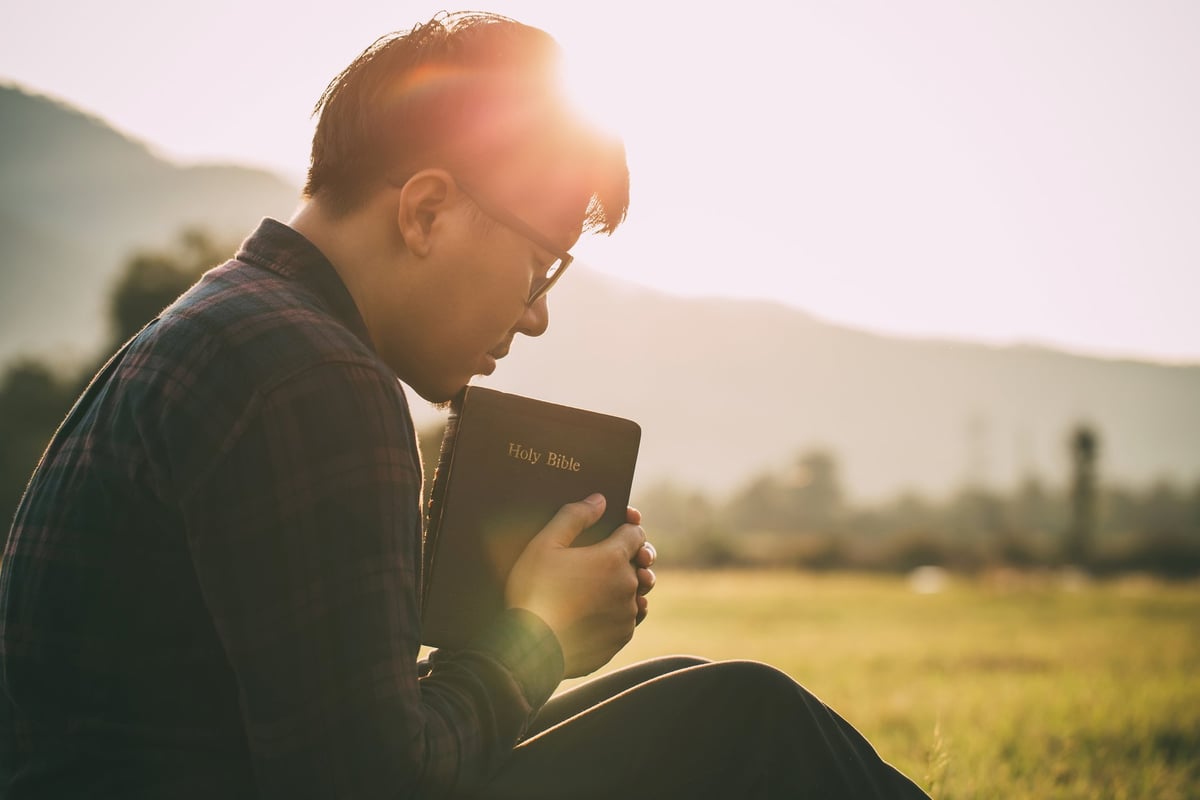 Man praying on the Bible in a field during a beautiful sunset