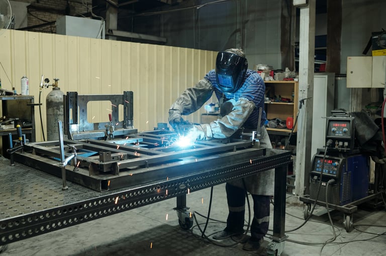 Young male worker of industrial plant bending over metallic workbench and welding parts of huge part of machine used in production