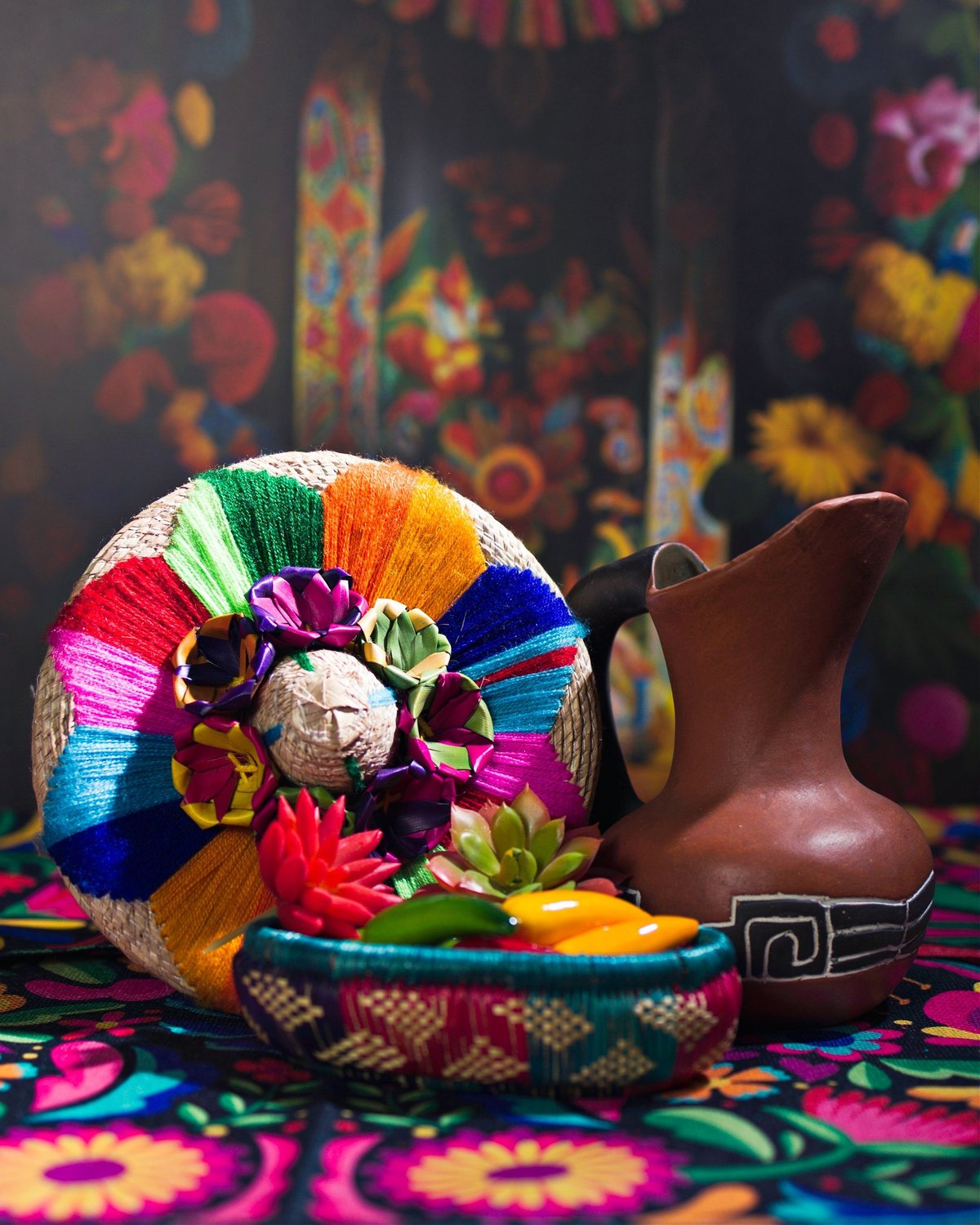 Traditional Mexican woven basket with chili peppers, avocado and clay jarritos jug on floral background