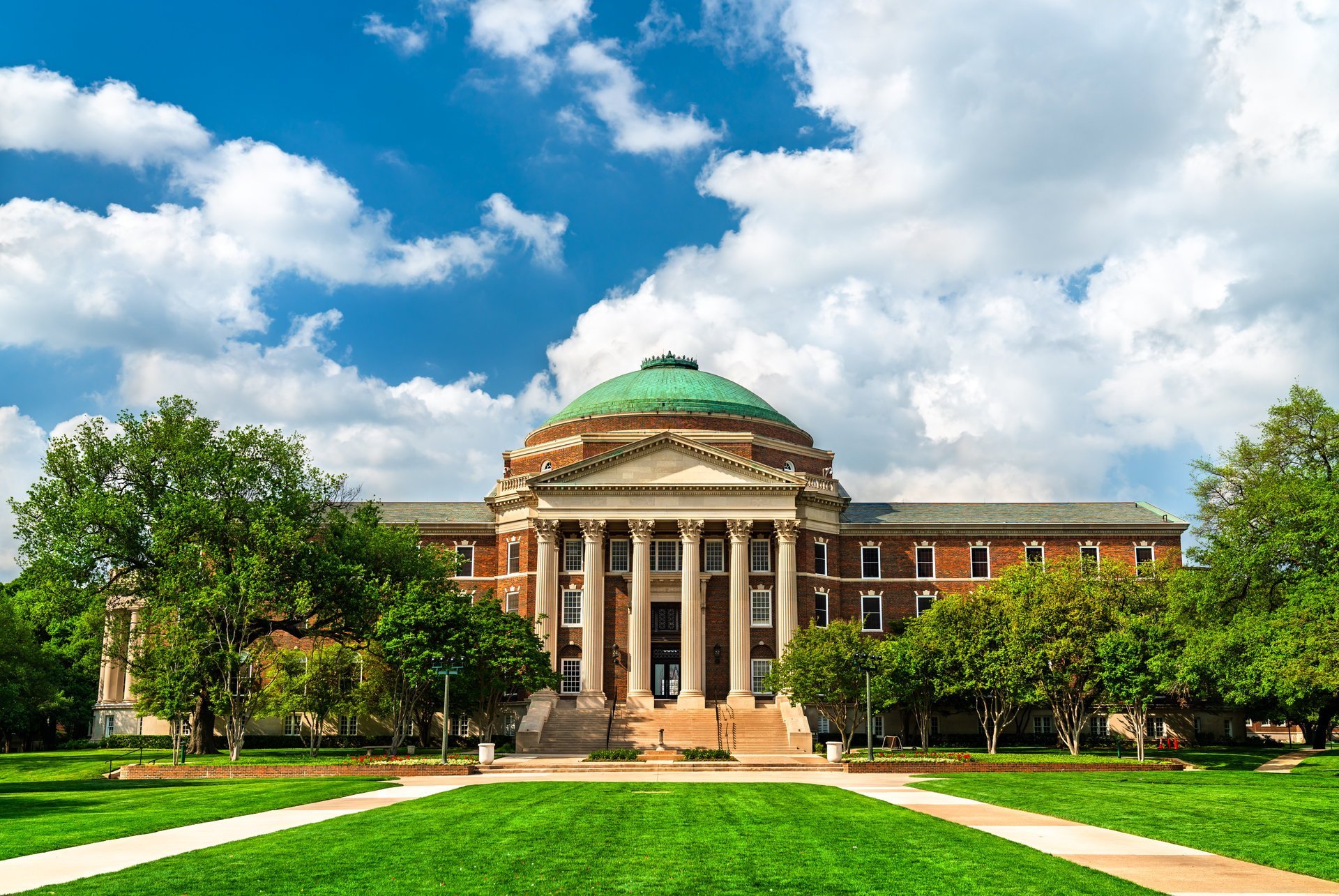 Historic university hall with classical columns, rotunda dome, and warm limestone facade set across a wide green campus lawn under bright daytime sky — independent campus housing information for TCU