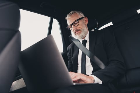 Senior businessman in suit working on laptop while being chauffeured in a luxury vehicle