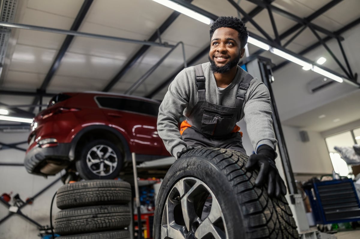 Portrait of hardworking smiling car technician rolling car tyre towards camera at automotive service center