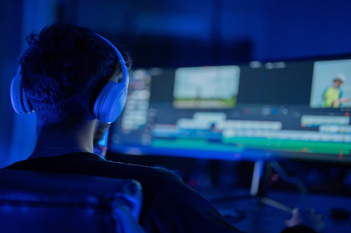A person with headphones engaged in video editing in a dimly lit room with a glowing screen showing clips and timeline