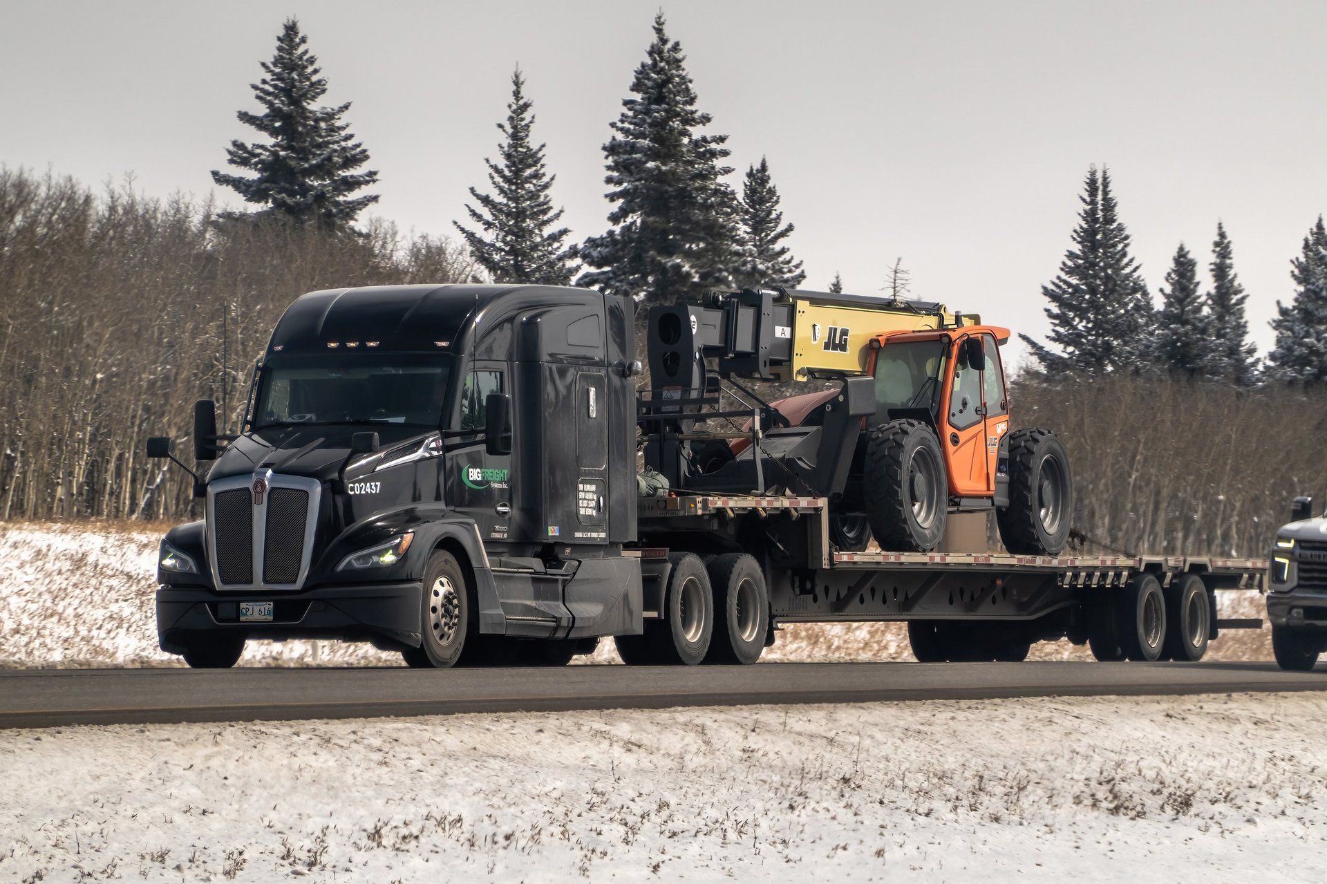 A black Kenworth T680 semi-truck hauling an orange JLG telehandler on a flatbed trailer along a snowy highway