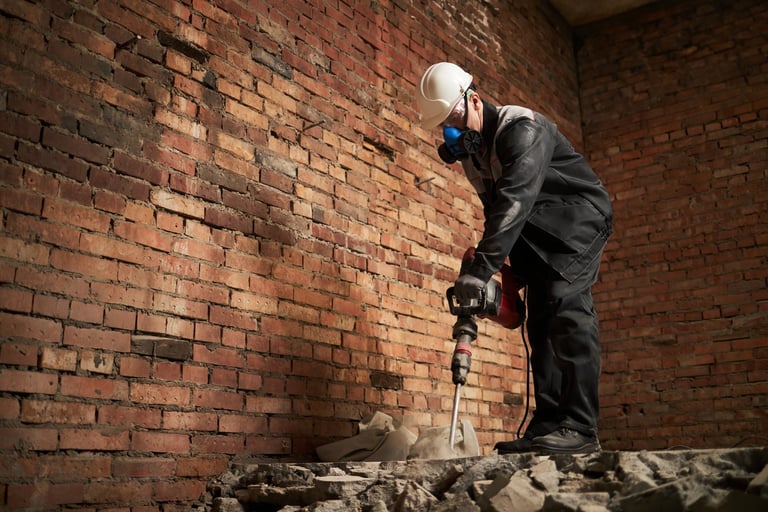 Caucasian man in dirty uniform wearing hardhat and respirator destroying cement floor with jackhammer