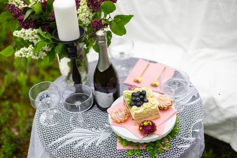 Romantic outdoor picnic table with lace, floral bouquet, champagne, and candle on a grassy field