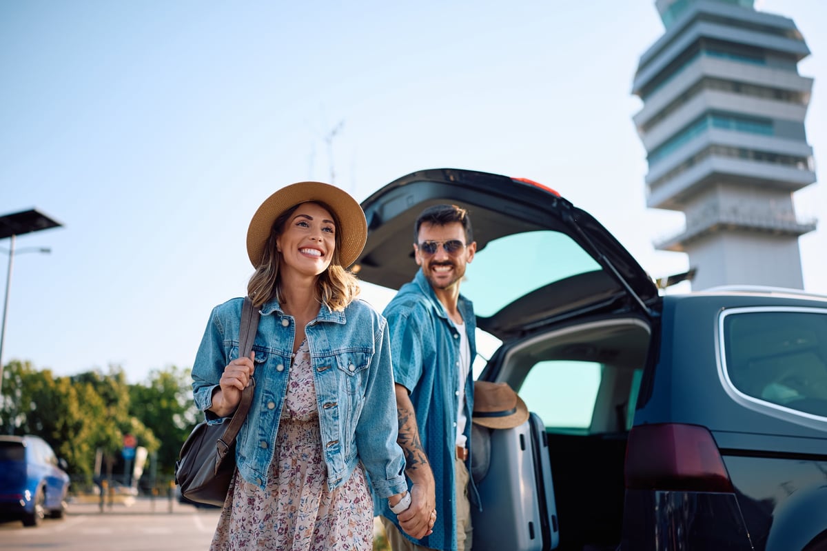 Travelers walking through an airport parking lot with luggage on a sunny day