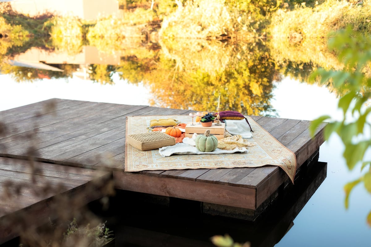 Cozy boho picnic with carpet, cushions, tray table, plaid blanket, and soft styled details near water