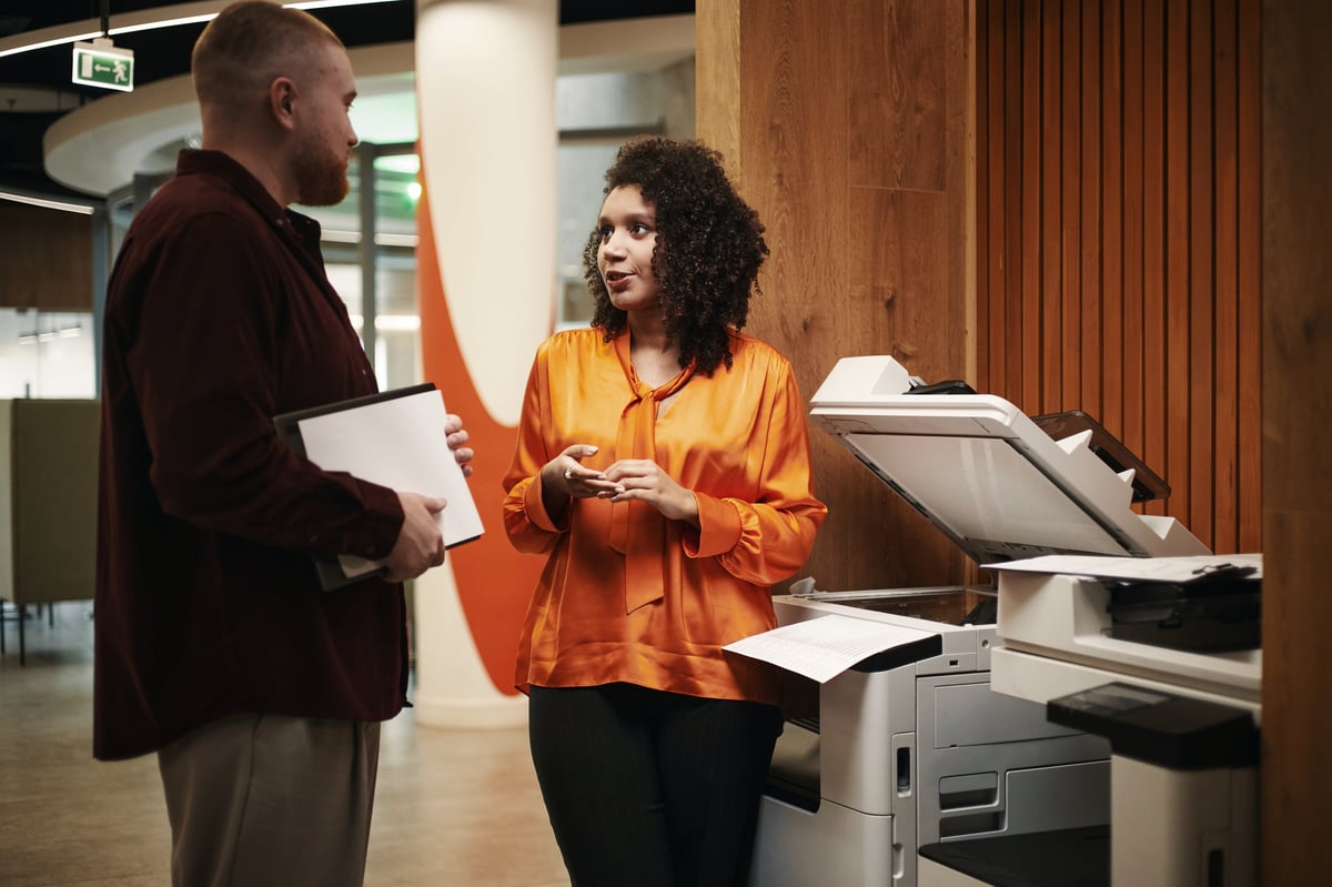 Employee receiving proactive technical support at a managed printer in the office while a colleague stands nearby with a folder