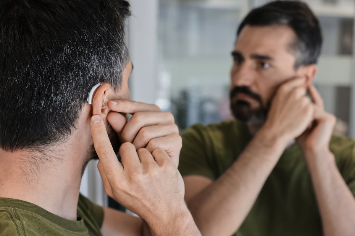 Adult man explores the comfort and fit of a new behind-the-ear hearing aid, adjusting it carefully in front of a mirror. Stylish hearing device