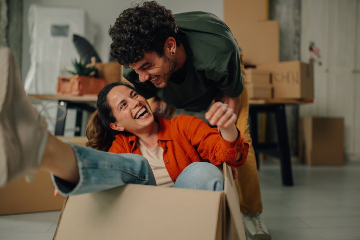 Young couple laughing and playing with a cardboard box while settling into their new apartment — the joy of moving forward