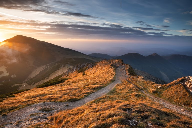 A mountain path stretching across a ridge at sunset, sky full of clouds and sunlight, distant peaks on the horizon