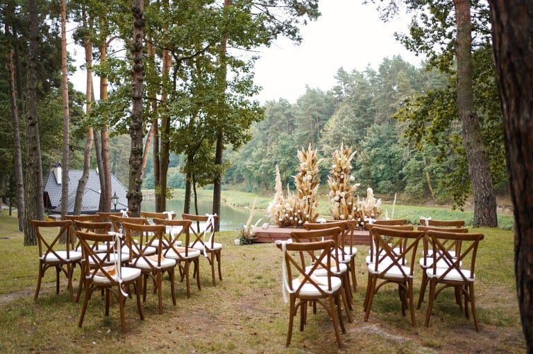 Rows of wooden chairs facing wedding arch with flowers and dried grasses in forest near the water