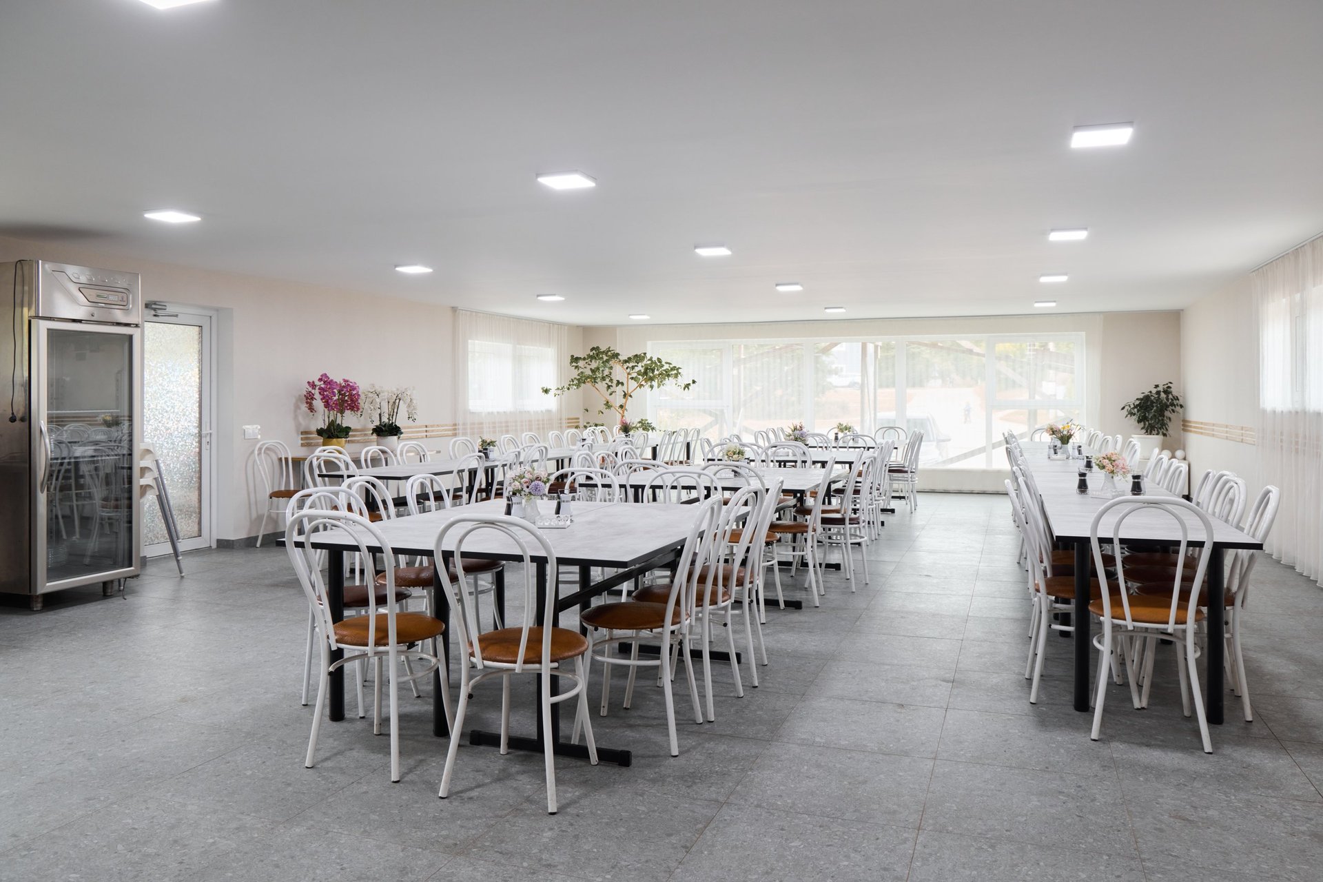Spacious event dining area with white chairs and wooden tables in bright natural light, cleaned and reset after a celebration