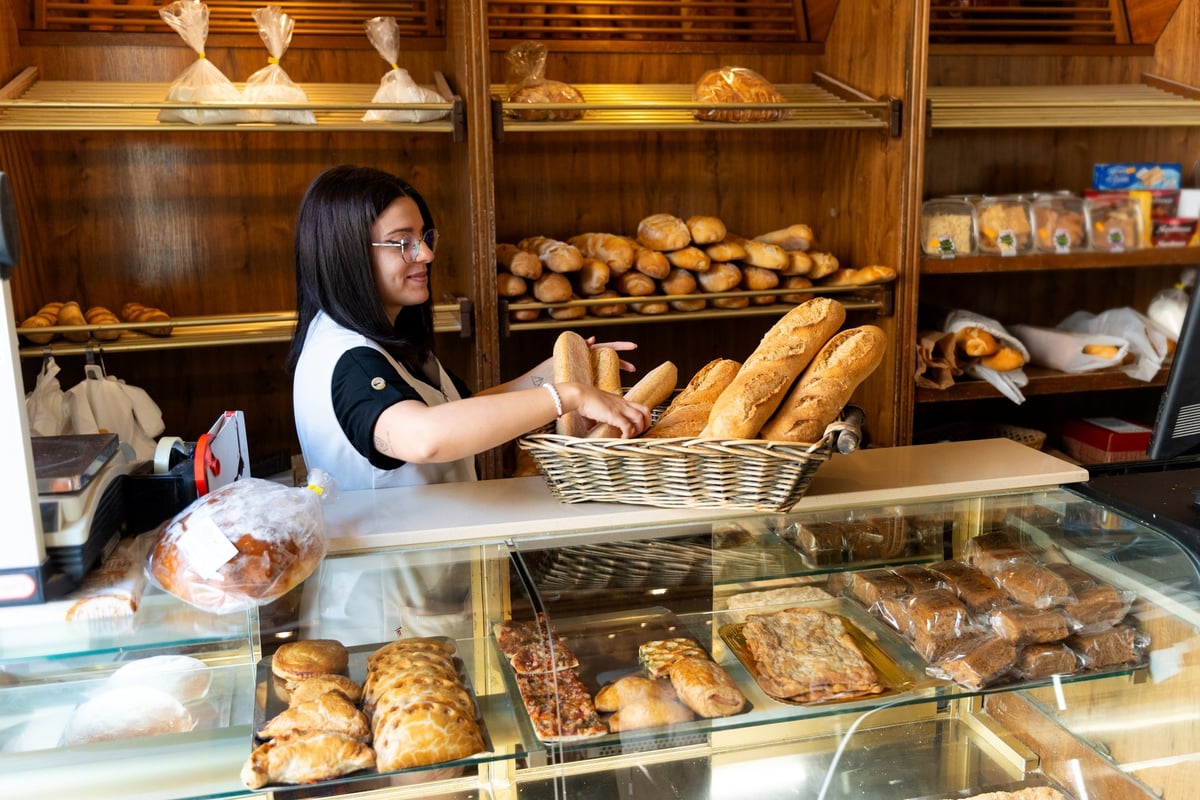 Young adult woman baker tidying up fresh baguettes in a woven basket on the counter of a small bakery shop