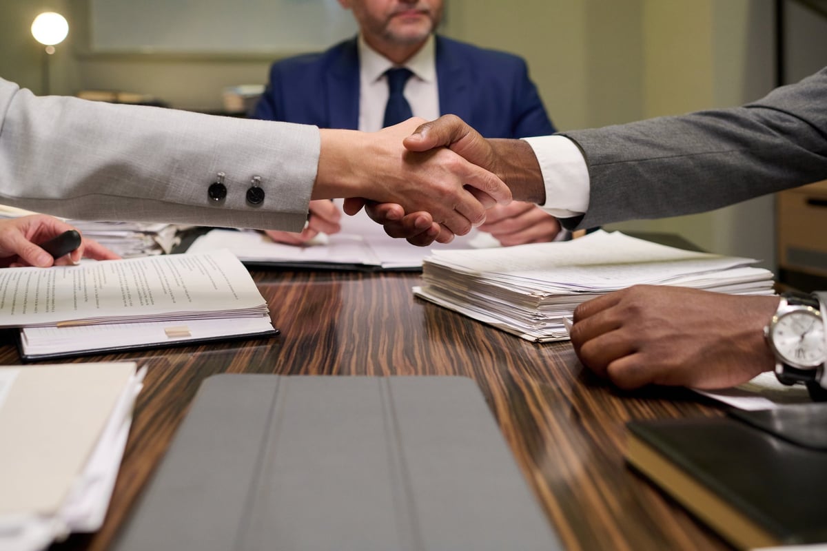 Caucasian woman and Black man shaking hands over stack of documents during business meeting, middle aged man in suit sitting in background, business contract negotiation scene