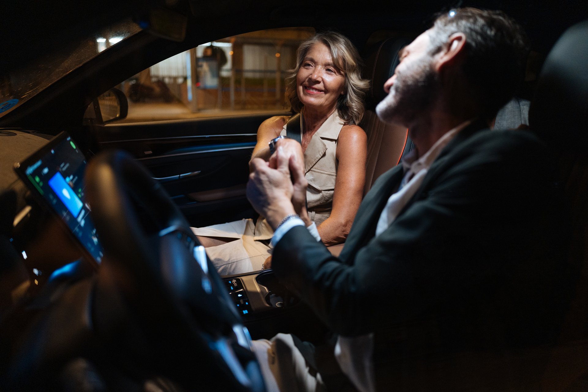 A smiling woman sits in the passenger seat while a man drives, sharing a light moment during a night ride through the city