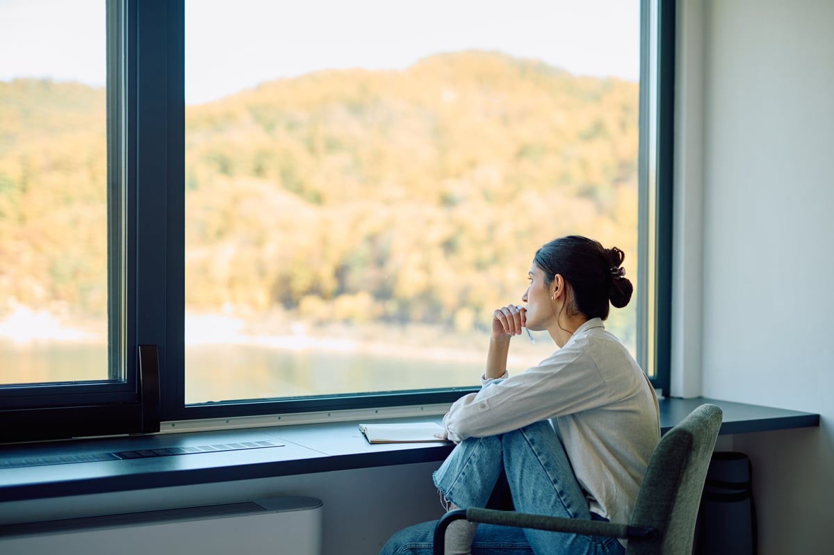 Young woman sitting by a large window, looking out thoughtfully — a quiet moment of imagining the next chapter
