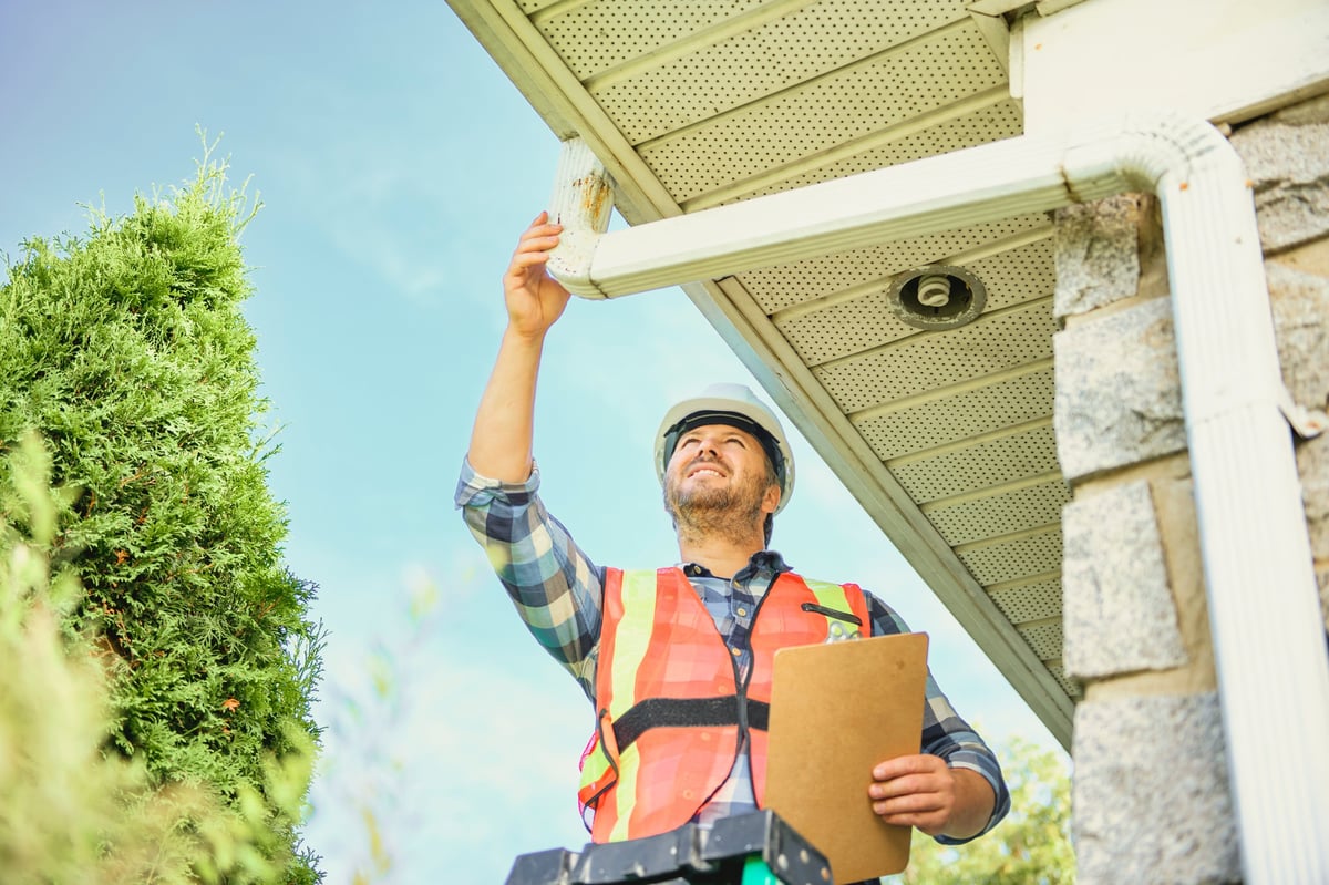 A man with hard hat standing on steps inspecting house gutter
