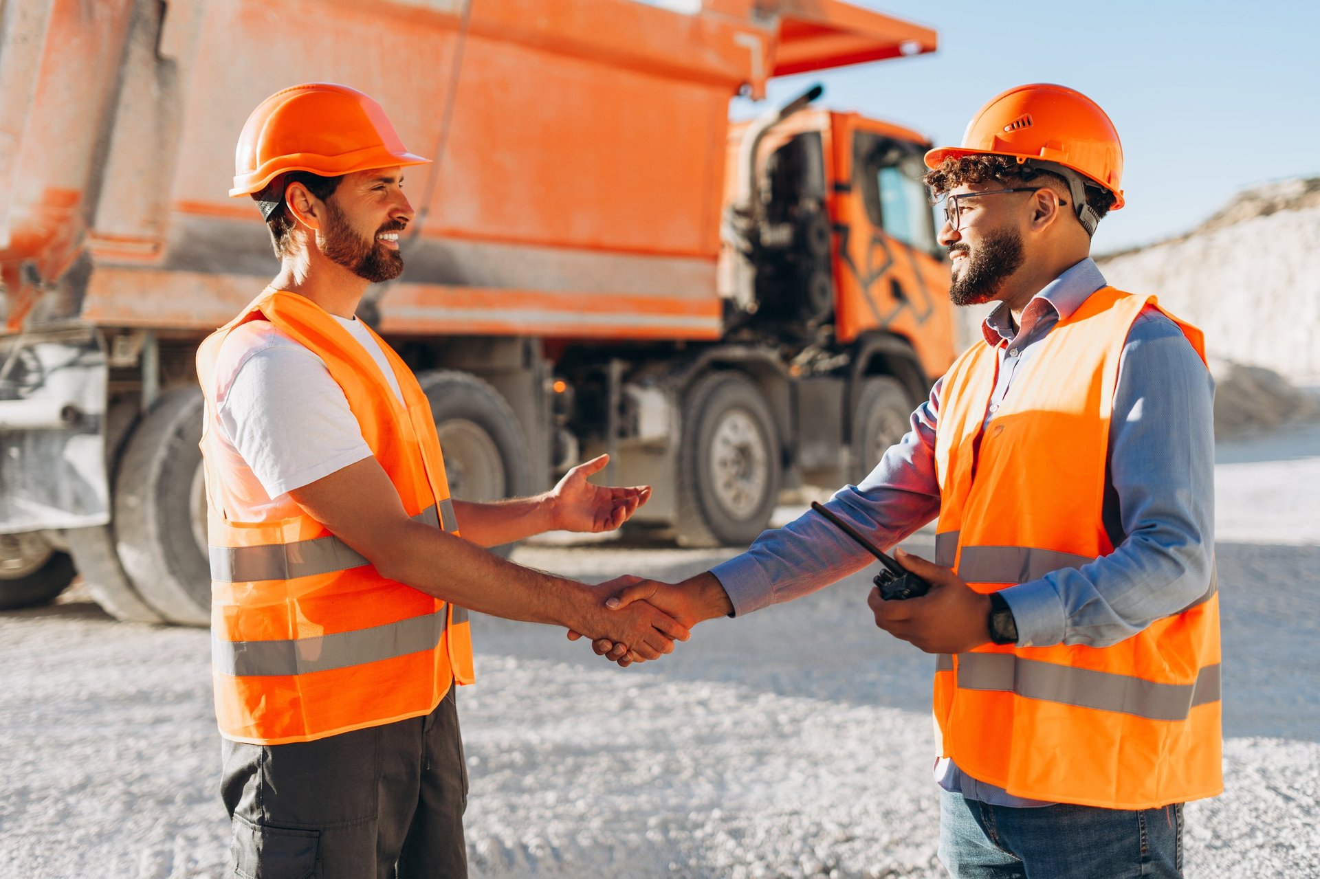 Professional team, smiling construction workers wearing orange hard hat and vests shaking hands at quarry site. Partnership, teamwork concept