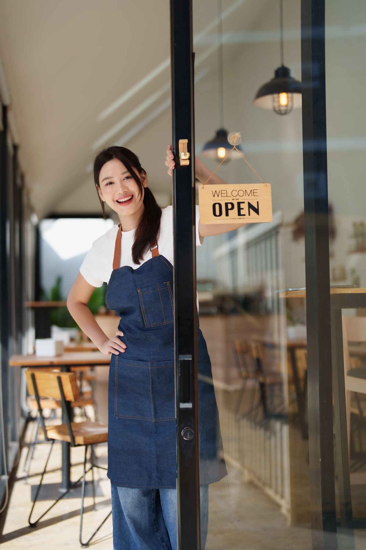 Young female cafe owner stands in her open doorway smiling and inviting customers into her modern small business