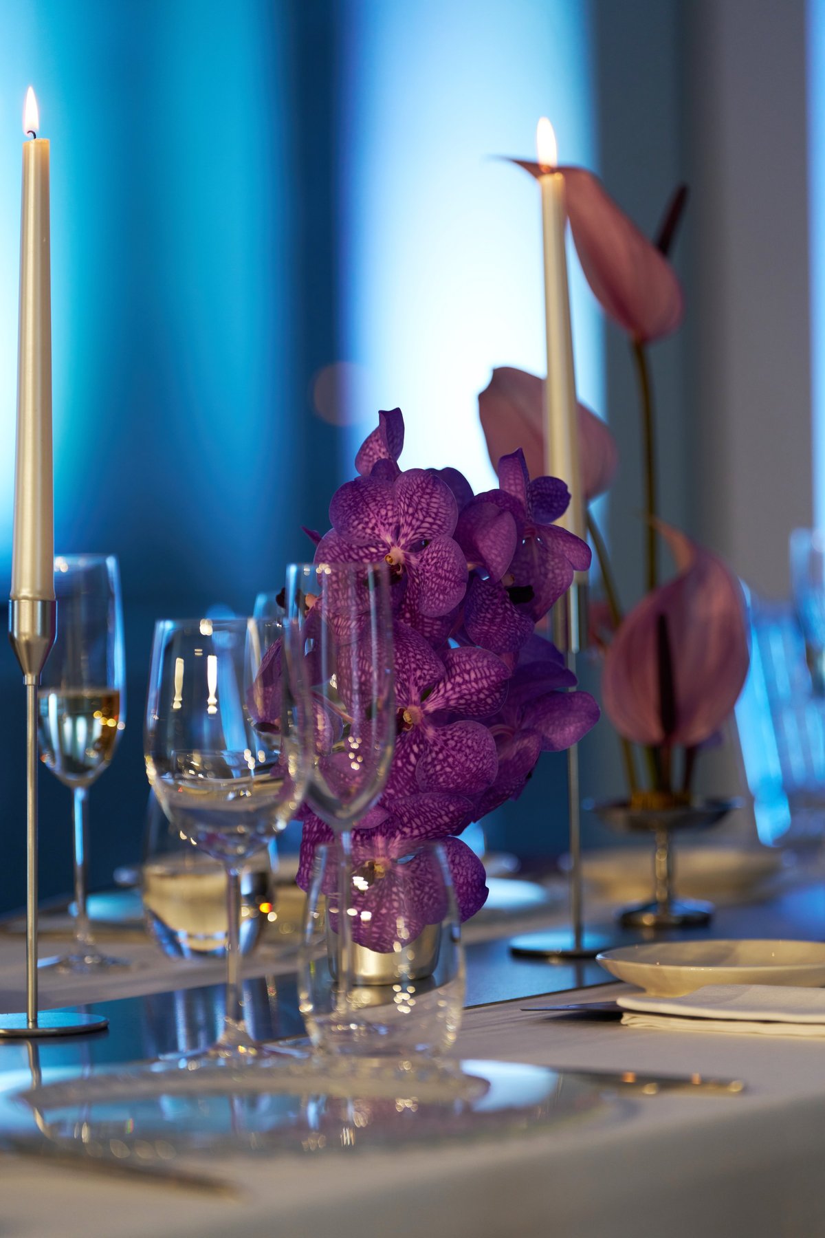 Elegant dinner table setting with crystal glasses, candles, and a centerpiece of vibrant purple orchids against a moody blue background