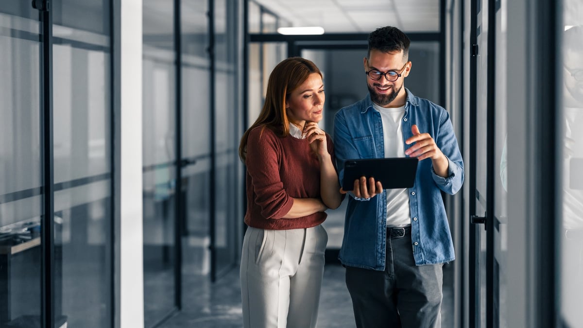 Two colleagues standing in a modern office hallway collaborate and discuss ideas using a tablet