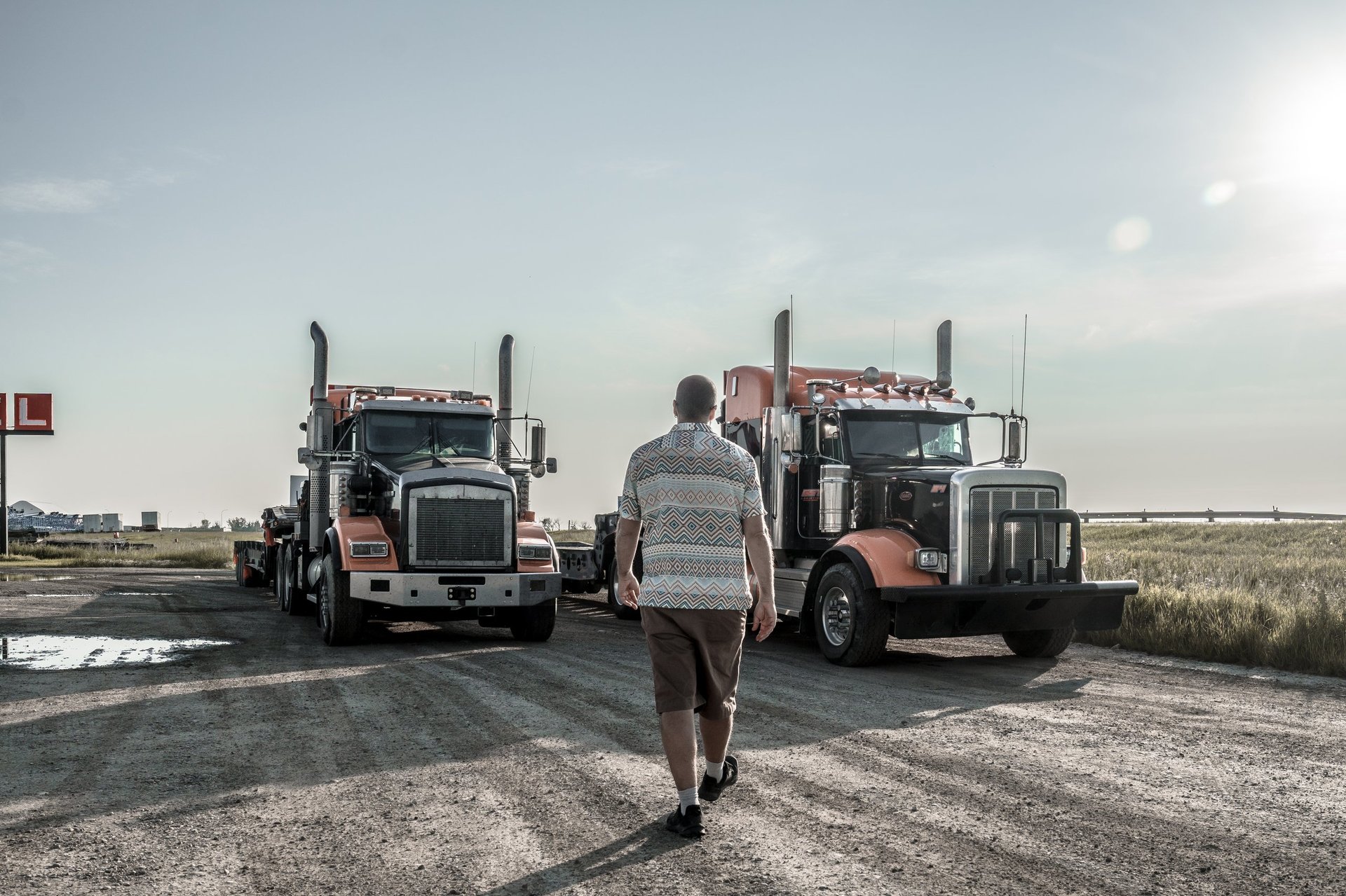 A trucker walking along a dirt road toward two semi-trucks parked near a field in the Canadian Rockies