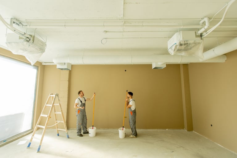 Two painters focused on painting a wall in a well-lit room using long rollers and paint buckets
