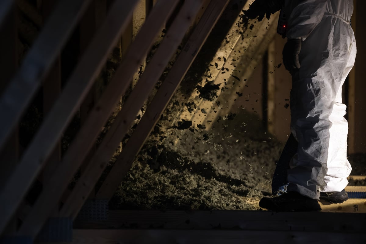 Worker in white suit blowing loose fill cellulose between wooden rafters in unfinished attic