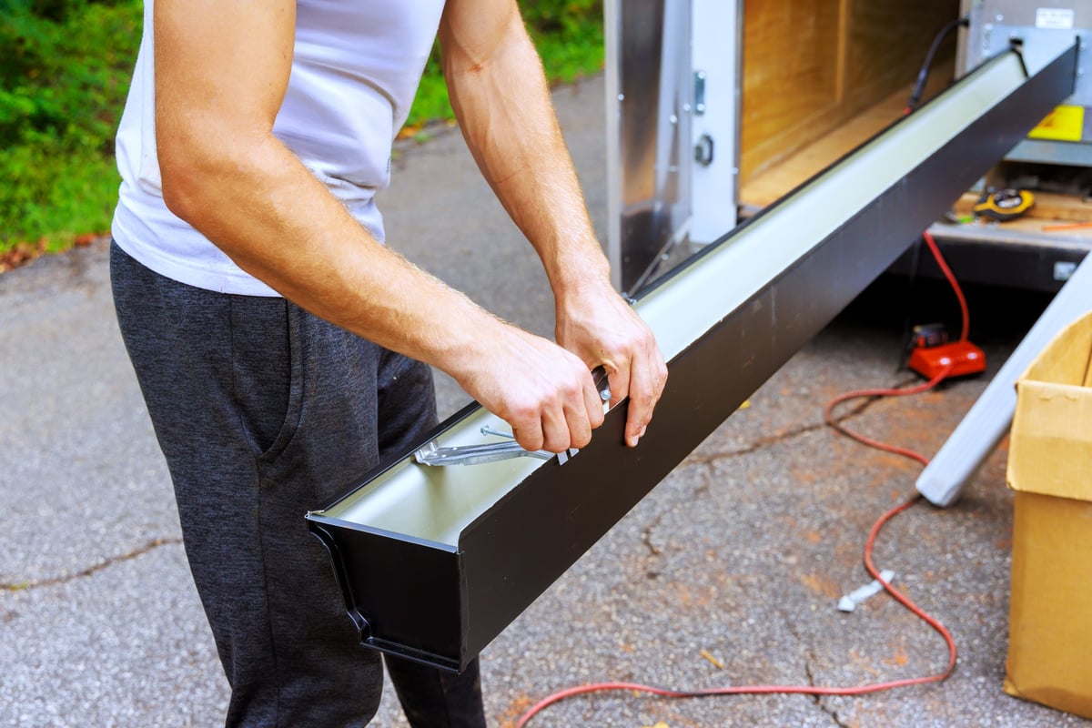 Man works on metal gutter channels with tool while near trailer outdoors