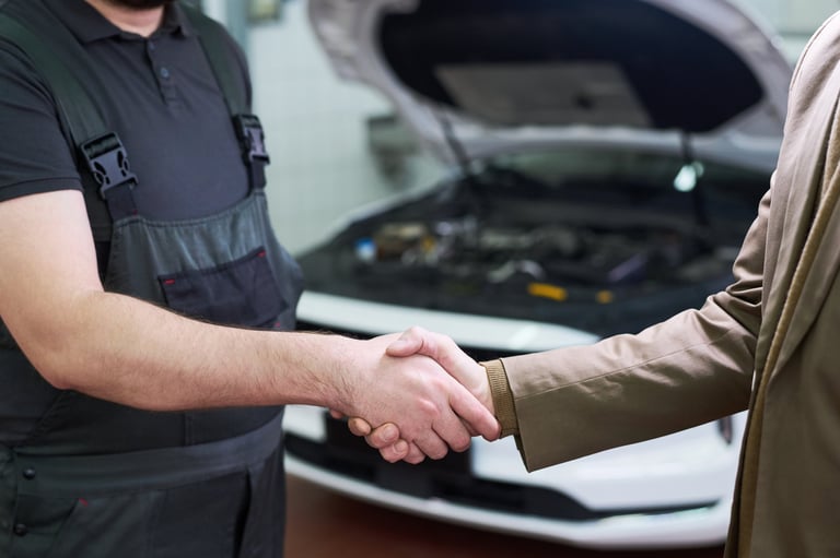 Man in work overalls shaking hands with client in suit in front of car with open hood in auto repair shop, confirming agreement