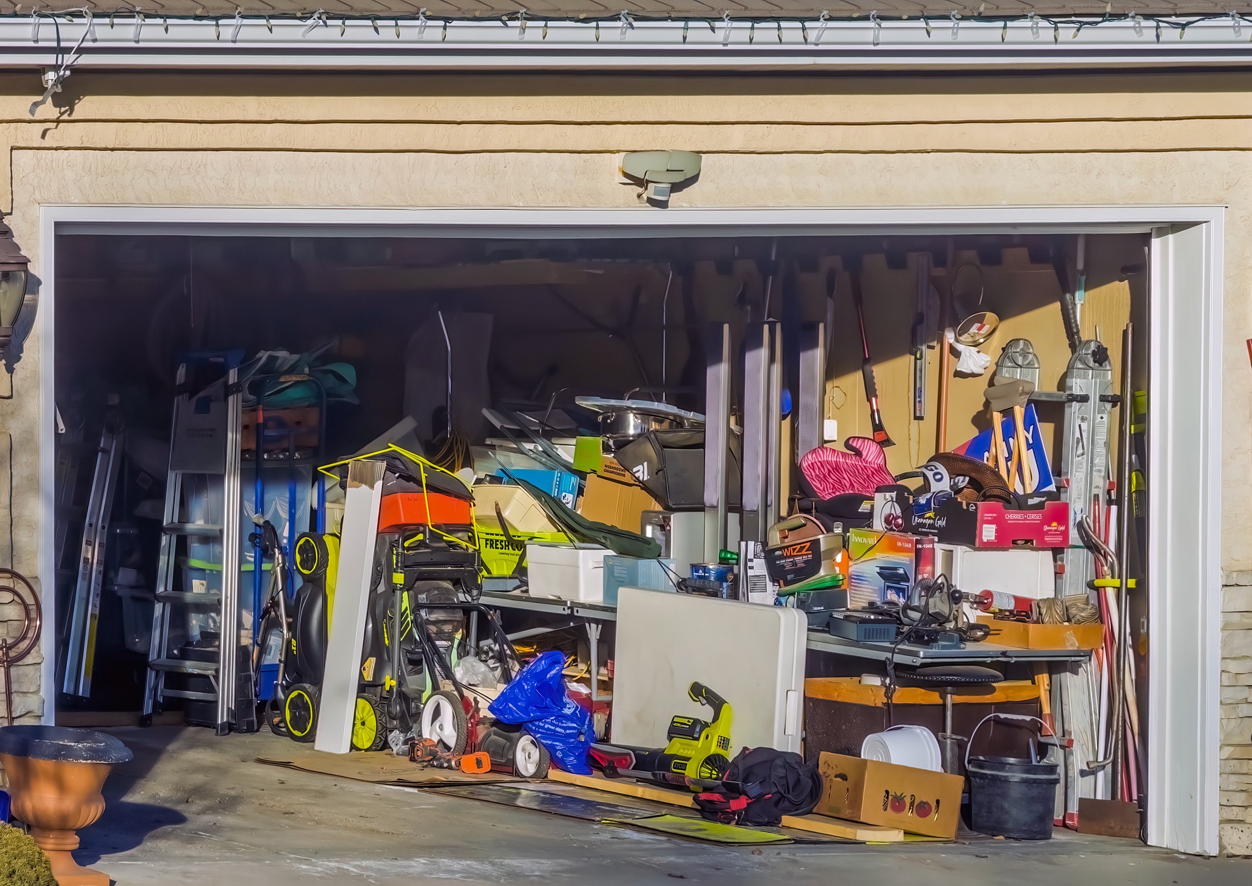 Cluttered suburban garage filled with overflowing household items, lawn equipment, tools and boxes piled up for a junk removal pickup