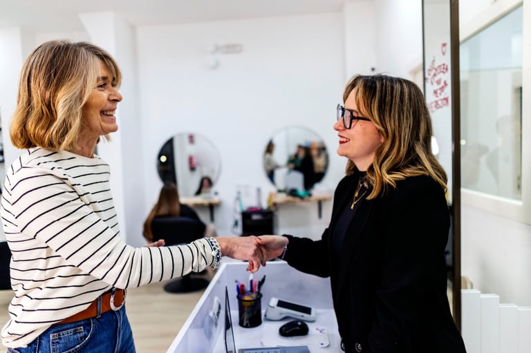 Two smiling women shaking hands at a hair salon reception desk