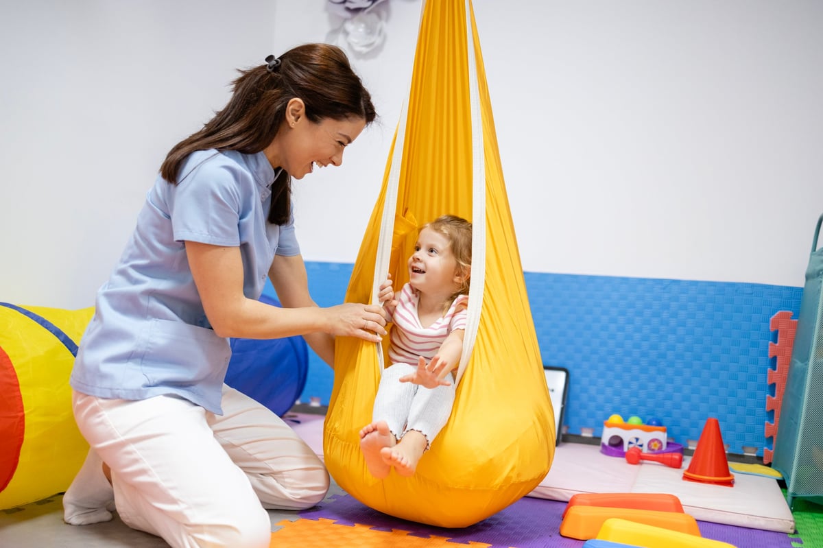 Female pediatric physiatrist assisting a young child in a sensory swing in a bright modern occupational therapy room