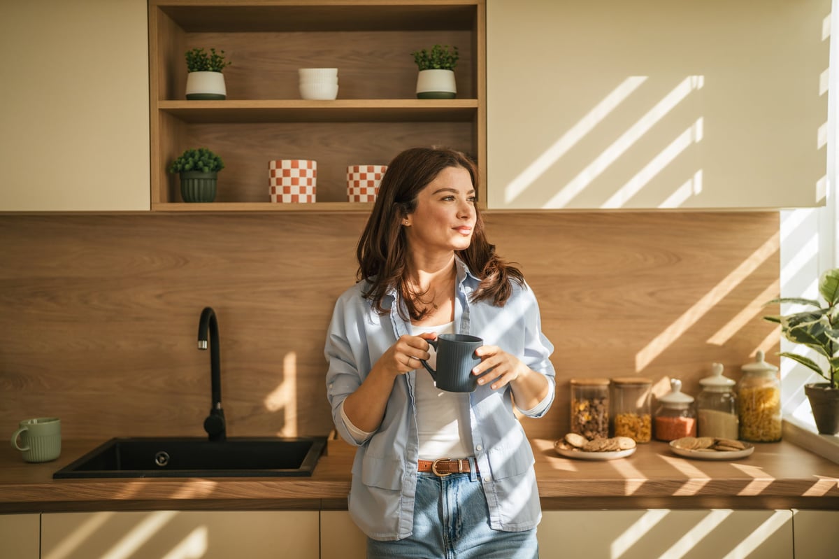 Woman standing in a modern kitchen holding a mug, illuminated by natural sunlight — a quiet, lived-in moment