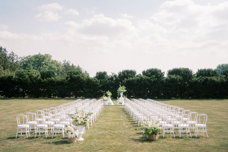 Wide outdoor wedding aisle lined with white chairs leading to floral arch, expansive lawn framed by trimmed hedges