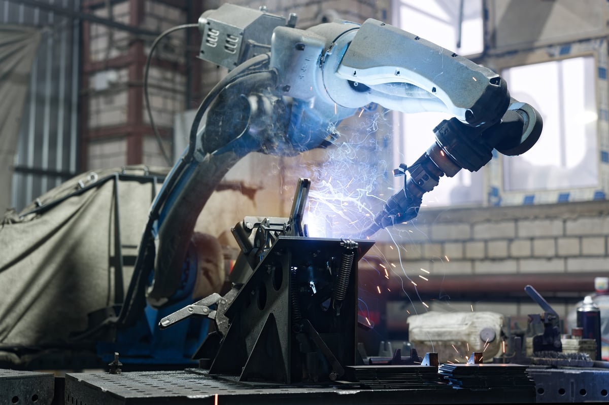 Robotic arm performing arc welding on a metal part secured with clamps, with sparks flying in a high-tech automated production environment