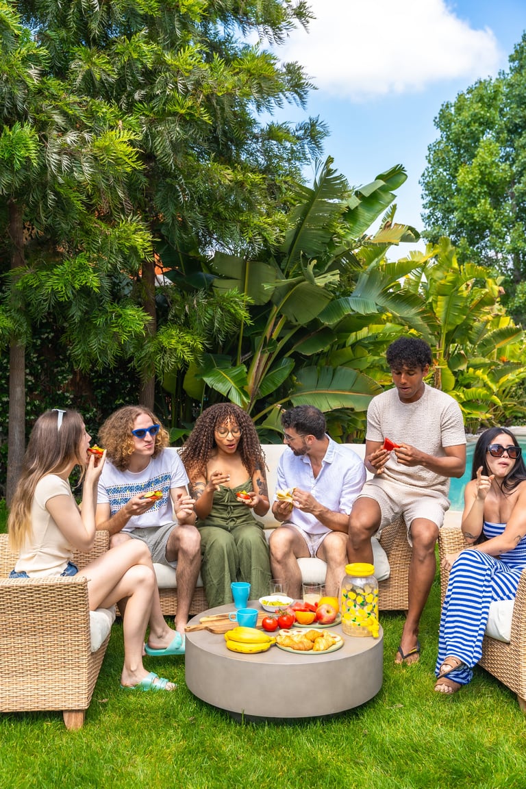 Group of diverse friends sitting on outdoor furniture in a lush garden, enjoying food and drinks at a daytime social gathering