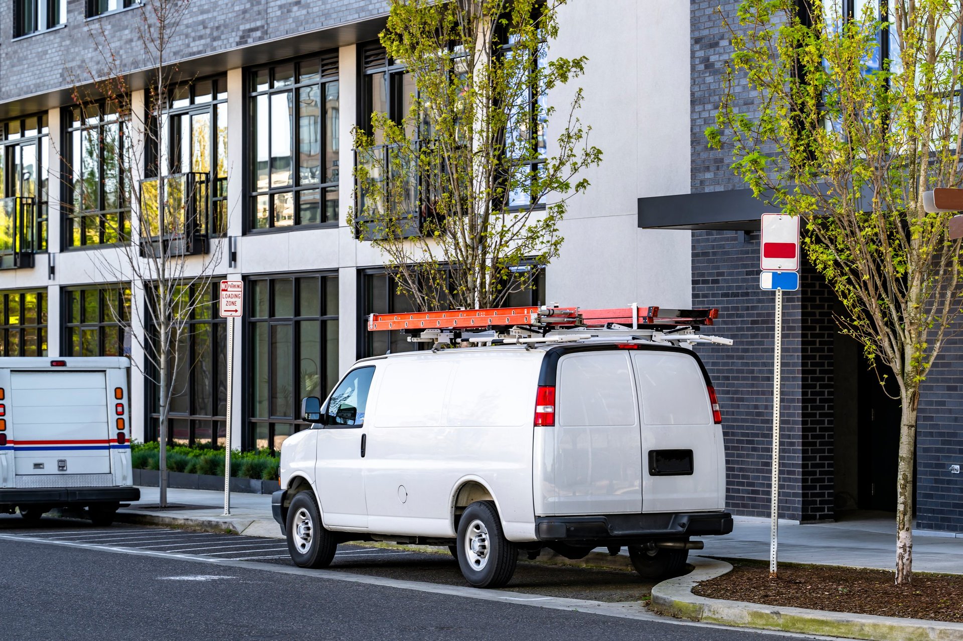 A white commercial cargo van with a ladder on its roof, parked on a city street