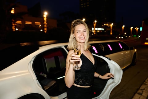 Young woman smiling while holding a glass of champagne, standing near a luxury limousine at night