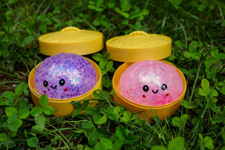 Two kawaii glitter squishy dumplings in purple and pink sitting in open yellow steamer baskets on grass