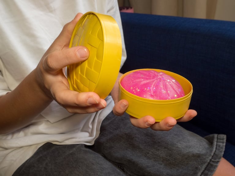 Close-up of a child's hands unboxing a pink glittery squishy dumpling from a yellow steamer basket