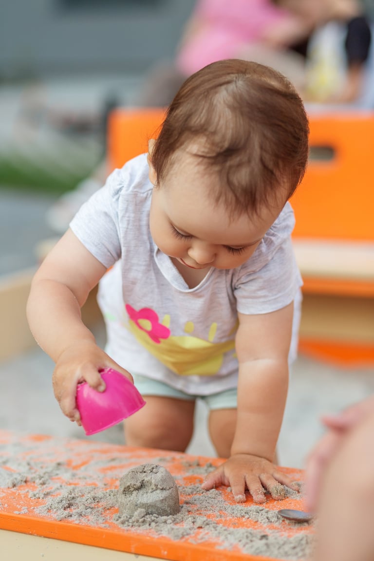 A toddler building sand shapes with a toy cup in an outdoor sandbox