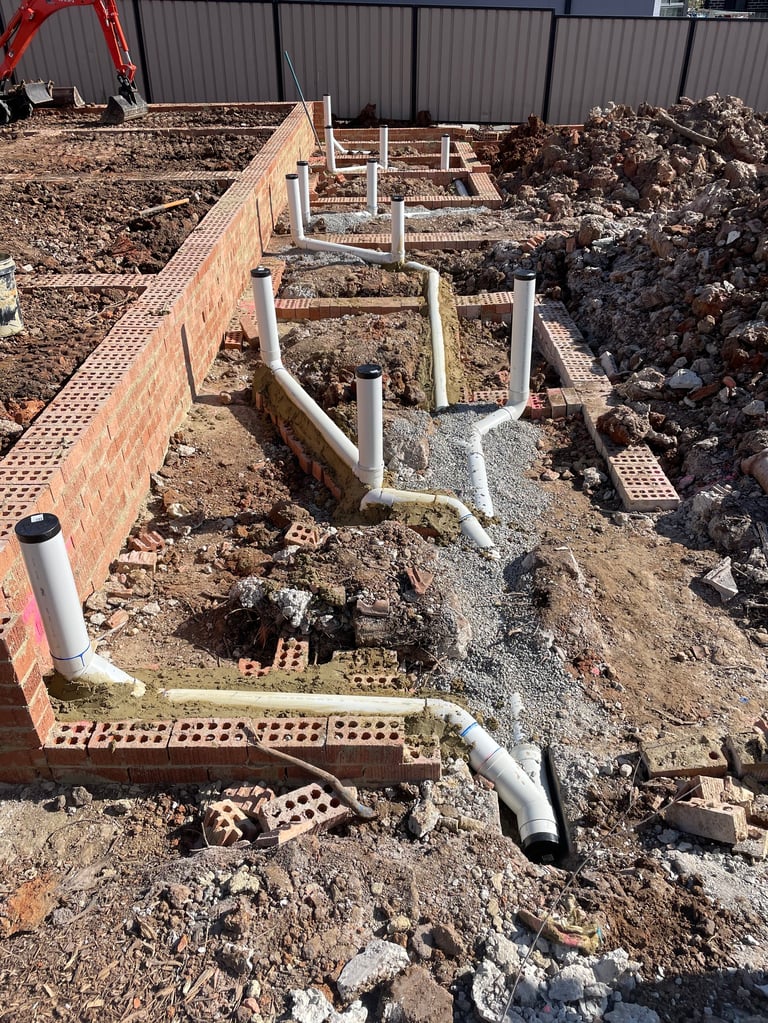 Construction site showing concrete foundation with brick walls, metal support posts, and drainage pipes laid in sandy soil