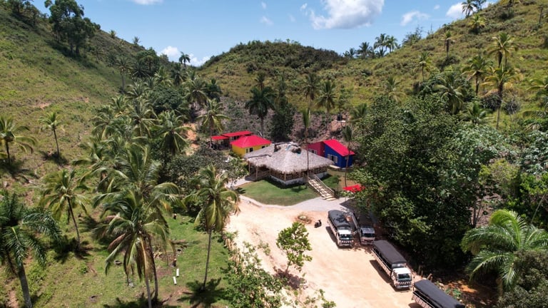 Aerial view of a glamping resort nestled in lush green hills with pink glamorous tents, palm trees, and a central pavilion area