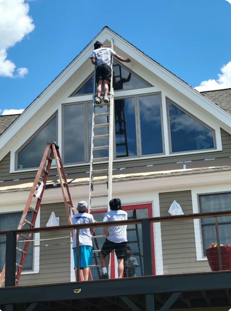Workers cleaning large gable windows on residential house exterior from ladder and scaffolding under blue sky