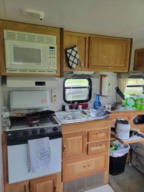 Before: RV kitchen with wood cabinets, microwave, electric stove, refrigerator, and countertops cluttered with cooking items and containers